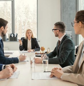 A woman in a business suit speaks to three colleagues during a meeting in a modern office. Documents and glasses of water are on the table, and large windows show trees outside.
