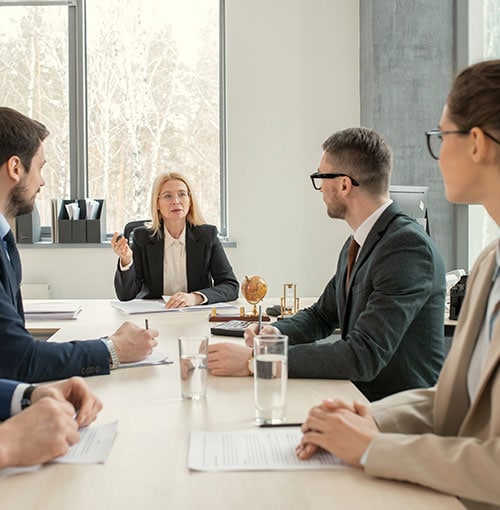 A woman in a business suit speaks to three colleagues during a meeting in a modern office. Documents and glasses of water are on the table, and large windows show trees outside.