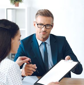 A man in a suit and glasses shows a clipboard with a blank sheet to a woman, who is holding a pen and looking at the paper, in a professional office setting.