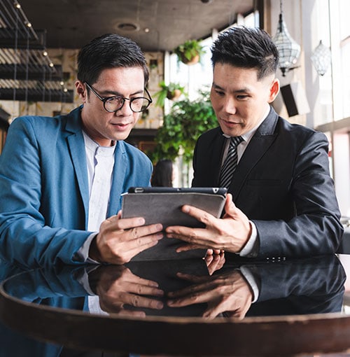 Two men in business attire looking at a tablet together while standing at a shiny table in a modern, well-lit office or café setting.