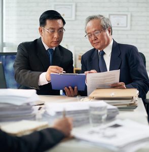 Two businessmen in suits sit at a desk, closely reviewing documents together. One holds a clipboard, while the other holds papers. The desk is covered with files and paperwork, and another person is blurred in the foreground.