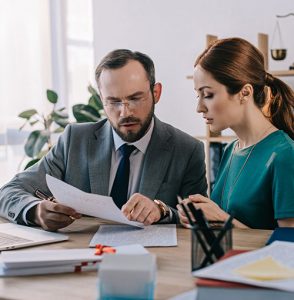 A man in a suit and glasses discusses a document with a woman in a green dress at an office desk, with papers, a laptop, and office supplies around them.
