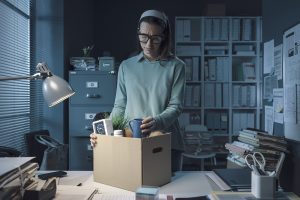 A woman in an office packs her belongings into a cardboard box, looking sad. The dimly lit, cluttered room hints she may be leaving due to wrongful termination NY, as paperwork and files are scattered around her desk.