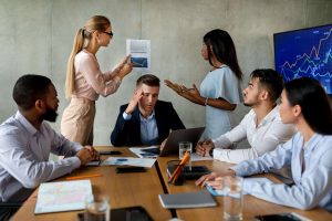 Six business professionals in a meeting room discuss charts and graphs, with one person holding their head in frustration while others converse and point at documents and a screen displaying financial data.