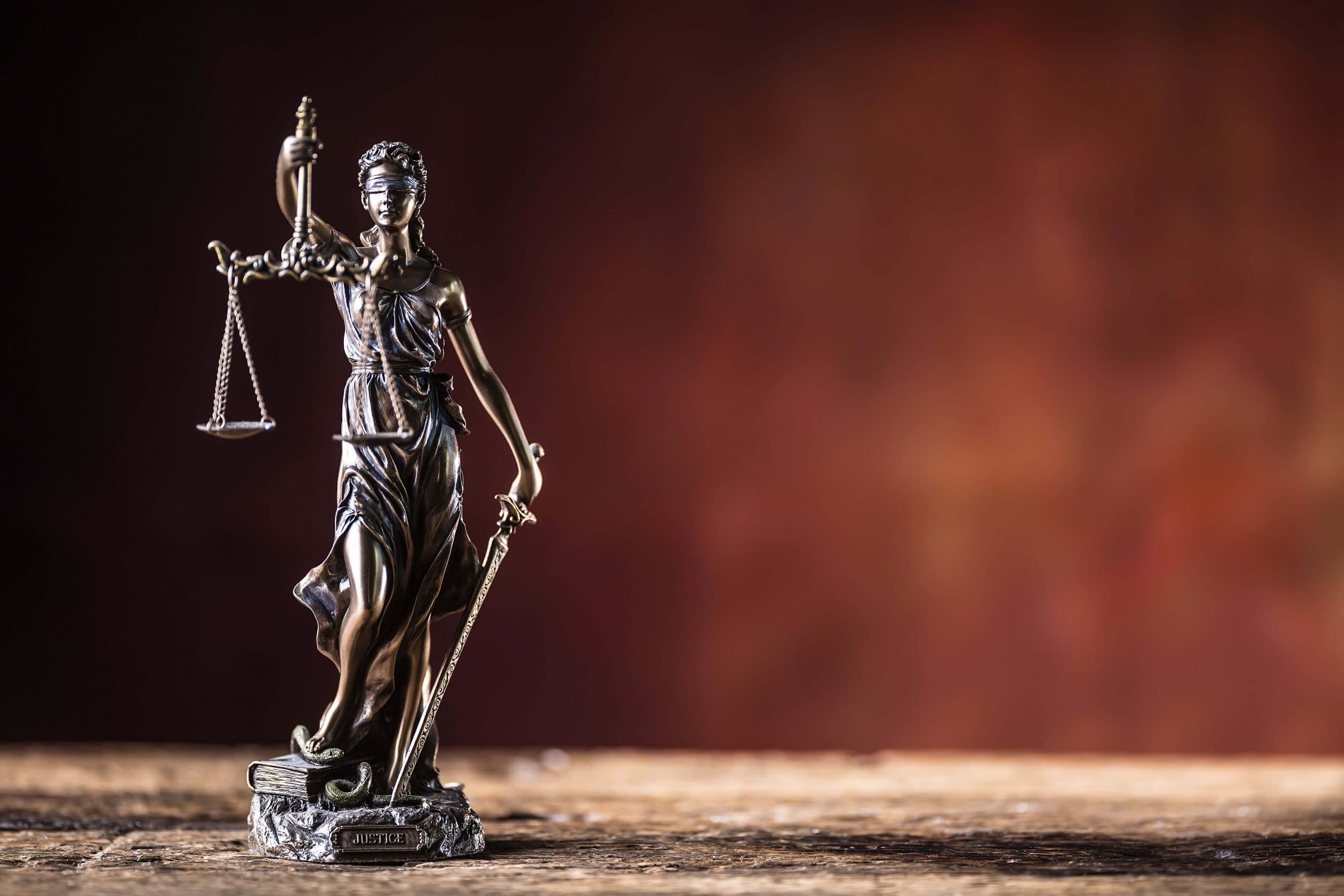 A bronze statue of Lady Justice holding scales in one hand and a sword in the other, wearing a blindfold, stands on a wooden surface with a blurred brown and red background.