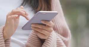 A person wearing a pink knit sweater uses a smartphone with a lavender case, touching the screen with one hand while holding the phone with the other. The background is blurred.
