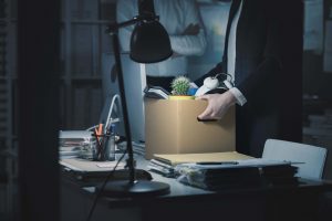 A person in business attire packs personal items, including a potted cactus and headphones, into a box on an office desk, suggesting they are leaving or being laid off. Another figure stands in the background with arms crossed.