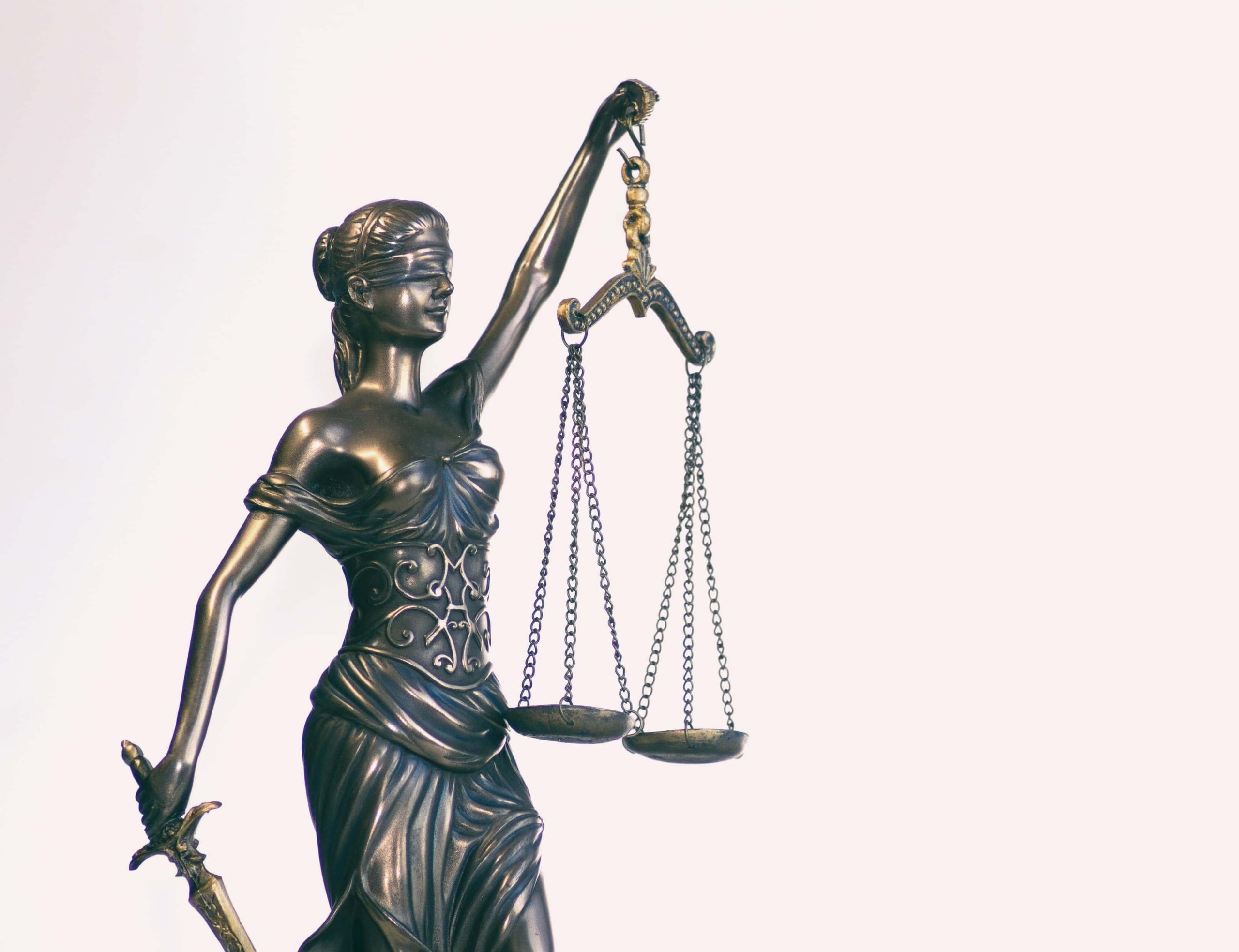 A bronze statue of Lady Justice holding a sword in one hand and balanced scales in the other. She is blindfolded, symbolizing impartiality, with a plain light background.