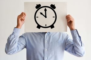 A person in a light blue shirt holds a paper with a large alarm clock drawing over their face, obscuring their identity. The clock shows the time as 8:00.