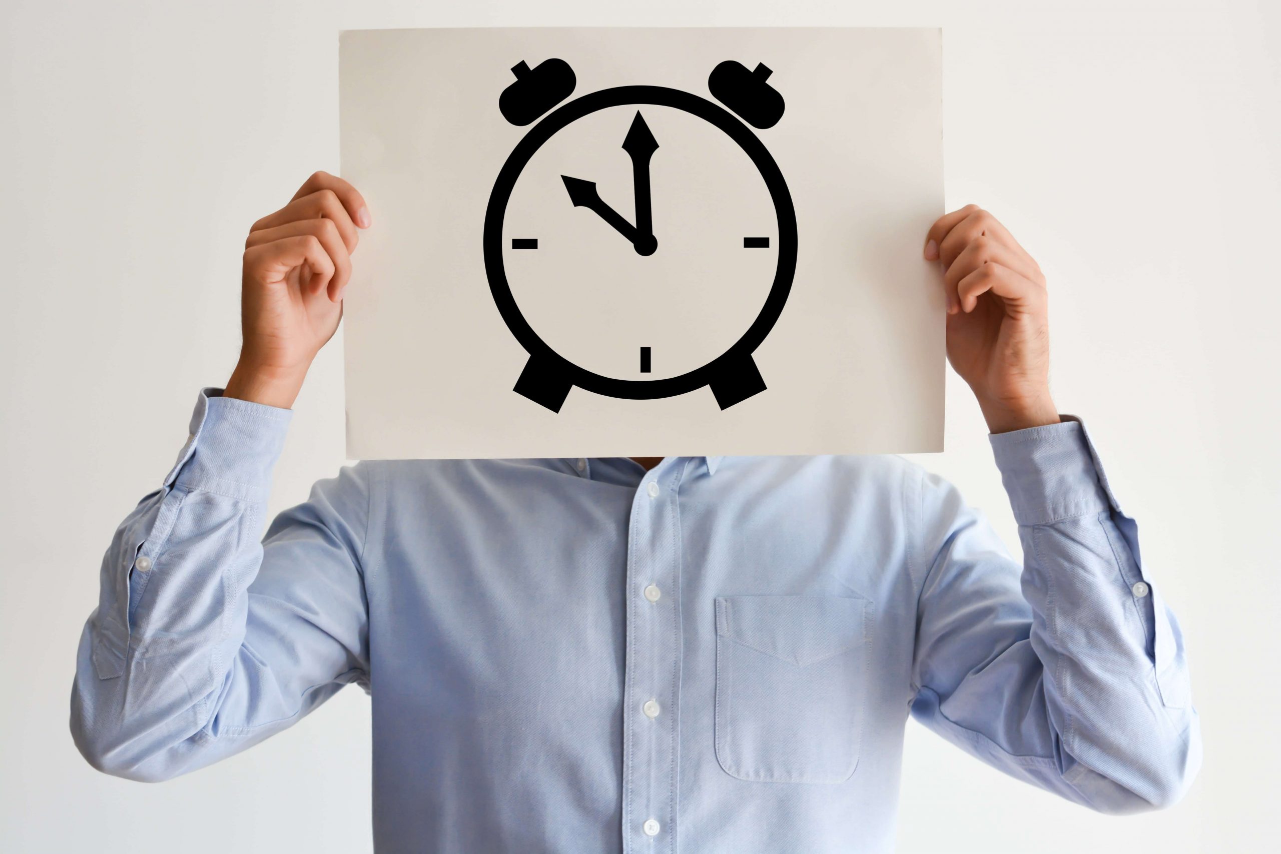 A person in a light blue shirt holds a paper with a large alarm clock drawing over their face, obscuring their identity. The clock shows the time as 8:00.