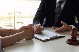 Two people sit at a desk discussing legal documents. One person gestures while the other holds a pen over a clipboard. A gavel and scales of justice are visible on the table, symbolizing a legal setting.