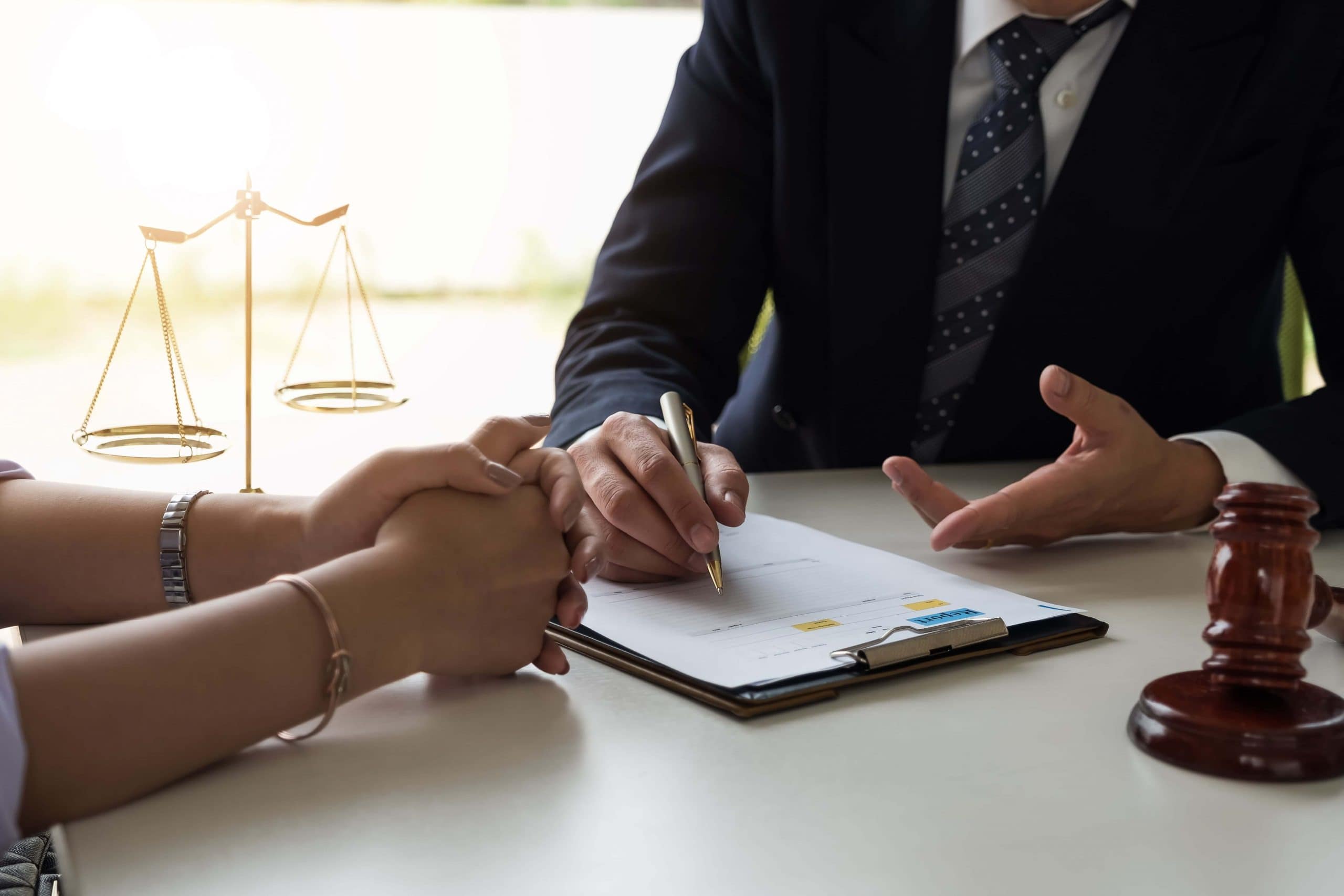 Two people sit at a desk discussing legal documents. One person gestures while the other holds a pen over a clipboard. A gavel and scales of justice are visible on the table, symbolizing a legal setting.