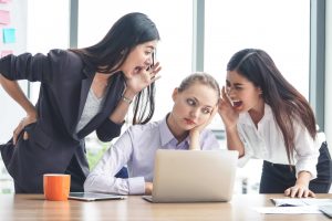 Two women are yelling at a third woman who looks frustrated and bored while sitting at a desk with a laptop. The scene takes place in an office with large windows in the background.