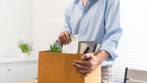 A person in a light blue shirt holds a cardboard box filled with office items, including a calculator, a small potted plant, and folders, suggesting they are packing up their desk, possibly after losing their job.