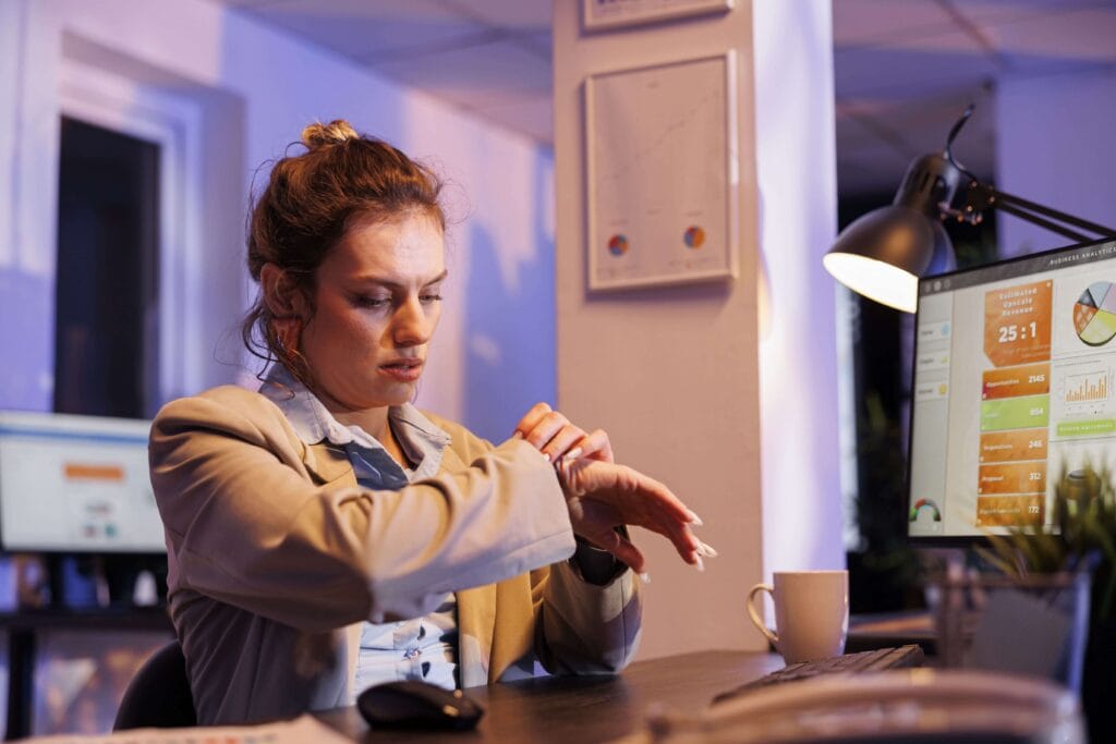 A woman in business attire sits at a desk in an office, looking at her watch. The desk has a computer monitor displaying charts and graphs, a lamp, and a coffee mug. The office appears to be dimly lit.