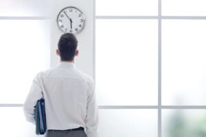 A man in a white shirt holding folders stands with his back to the camera, looking up at a clock on a white wall. The clock shows 9:00 as bright light streams through large windows—perhaps he’s considering advice from an overtime attorney.