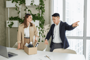 A woman stands by a table with a box of personal items, looking at a man pointing toward the door, suggesting she is facing whistleblower retaliation. Plants and large windows accent the modern office space.