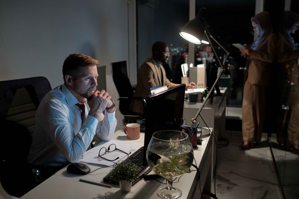 A man in a white shirt sits at a desk, looking thoughtful while working late in a dimly lit office. Two other people work in the background, perhaps consulting wage theft attorneys or reviewing documents by the window.