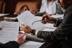 Close-up of people in business attire sitting at a table, holding and discussing printed documents related to a qui tam lawsuit, with one person holding glasses and gesturing toward the papers in a formal meeting or office setting.