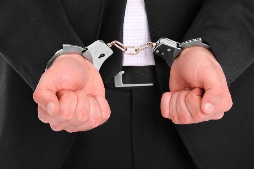 A person in a business suit with hands cuffed in metal handcuffs, shown from the front with their wrists held together, symbolizes the risks of whistleblowing and retaliation in the workplace.