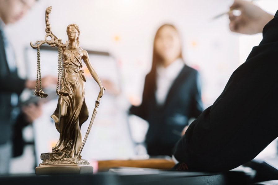 A bronze Lady Justice statue stands on a desk, with blurred business professionals in suits discussing sexual harassment in the workplace, symbolizing law and justice in a professional office setting.