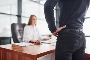 A person stands with a hand on their lower back in front of a desk, while a woman sitting at the desk looks at a computer—capturing a moment that could indicate concerns about sexual harassment in the workplace.