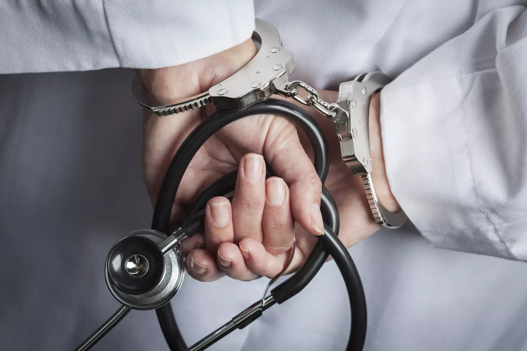 A person in a white coat is handcuffed behind their back, holding a stethoscope—hinting at the arrest of a medical professional, possibly linked to healthcare fraud whistleblower reward cases.
