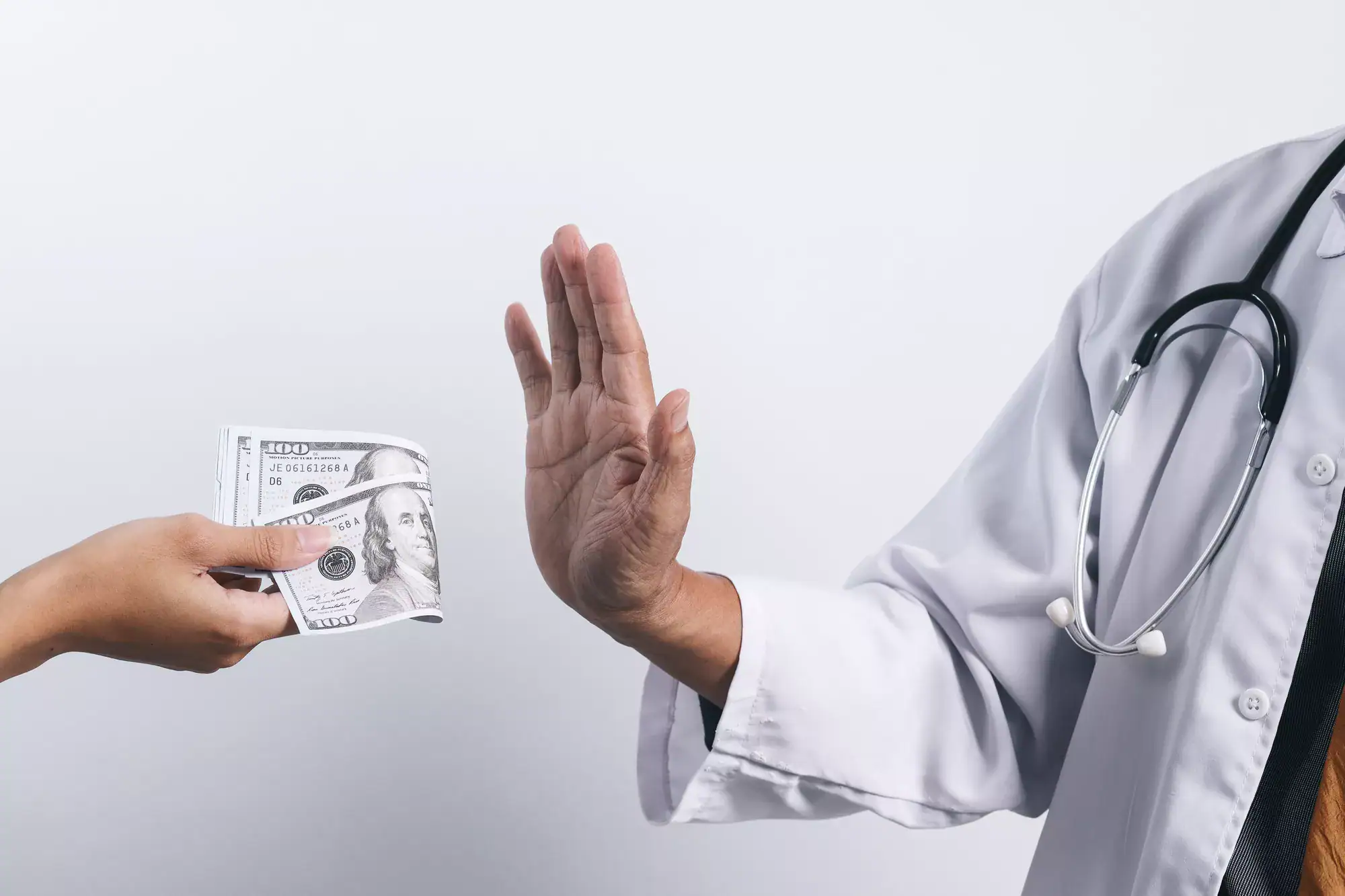 A person in a white coat and stethoscope raises a hand to refuse cash offered by another, highlighting the rejection of types of healthcare fraud or unethical payments in the medical field.