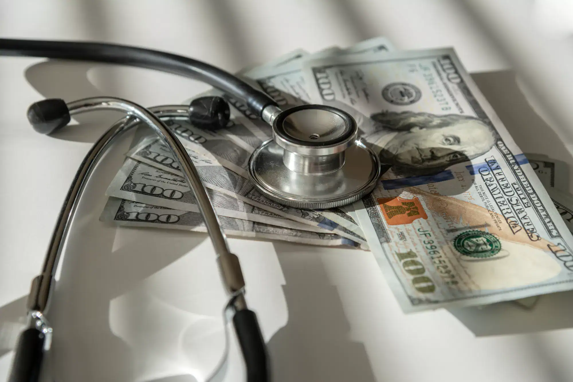 A stethoscope rests on US hundred-dollar bills, symbolizing the link between healthcare costs and financial incentives like a healthcare fraud whistleblower reward. Shadows and light create a dramatic effect on the white surface.