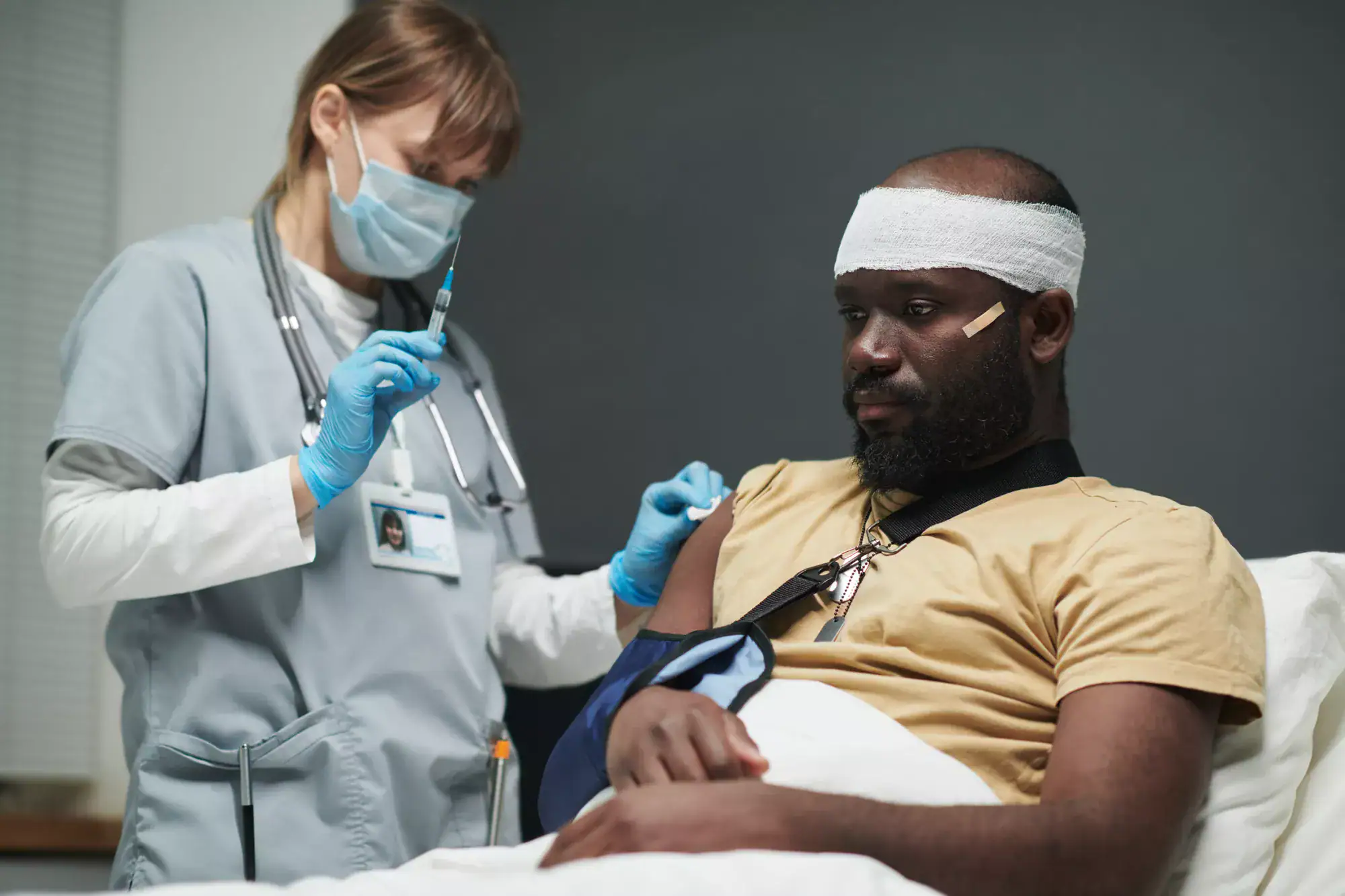 A nurse wearing gloves and a mask gives an injection to a man with a bandaged head and arm sling, sitting calmly in a hospital bed. The scene highlights patient care, contrasting genuine treatment with the dangers of certain types of healthcare fraud.