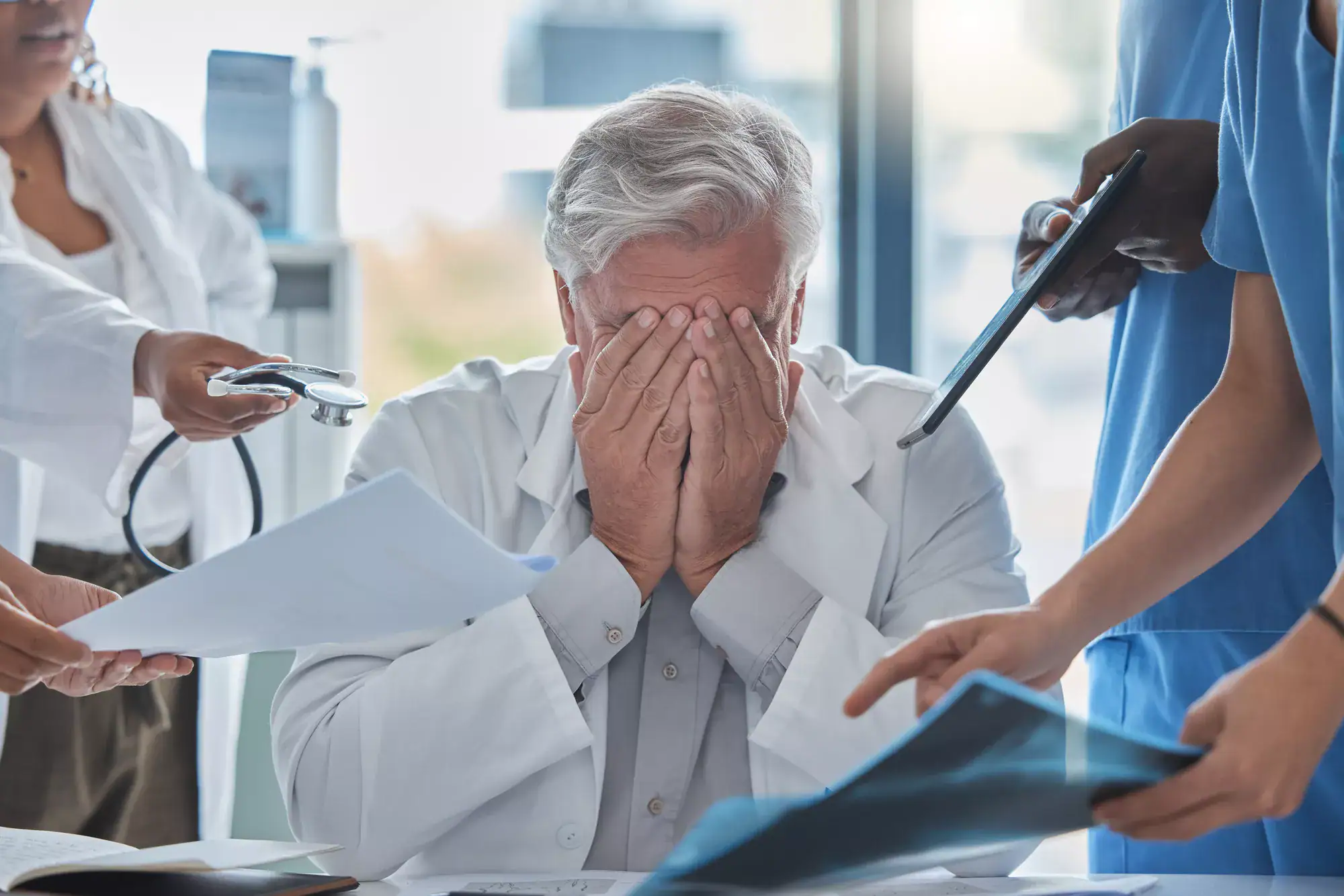 A doctor sits at a desk with his face in his hands, looking stressed, while several people around him hand him medical charts and documents—possibly overwhelmed by the complexities of different types of healthcare fraud.