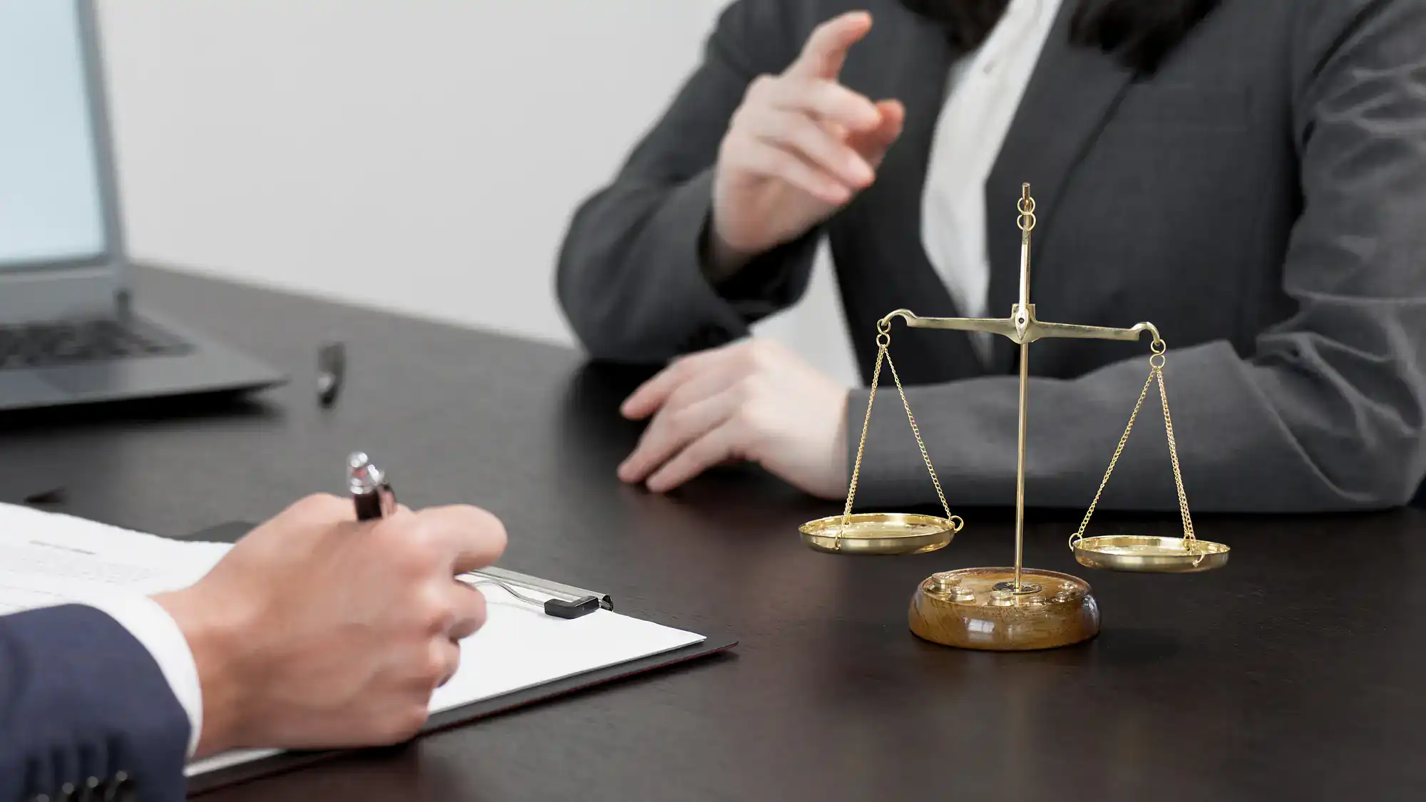 Two people in business attire sit at a desk with a laptop, clipboard, and golden balance scale, suggesting a legal consultation&mdash;perhaps with a sexual harassment attorney Manhattan trusts for expert guidance on NY workplace issues.