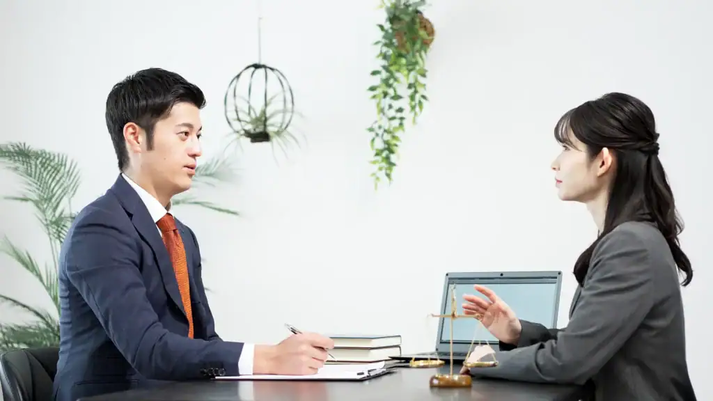 Two people in business attire sit across from each other at a desk in a NY office, engaged in conversation. A laptop, books, and a small scale are on the table—ideal for a sexual harassment attorney Manhattan consultation. Plants hang on the white wall behind them.