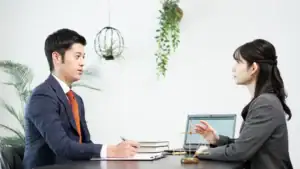 A man and a woman in business attire sit across from each other at a desk, discussing. The woman gestures with her hand as they consult a sexual harassment attorney Manhattan. A laptop, books, golden scales, and green plants complete the scene.
