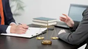 Two people in business suits sit at a desk with a scale of justice, a notebook, and a laptop, suggesting a legal or professional meeting. One person writes while the other gestures with their hand.