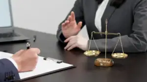 Two people in business attire sit at a desk with a scale of justice, clipboard, and laptop, suggesting a legal discussion. One appears to be an NY sexual harassment attorney in Manhattan meeting with a client.