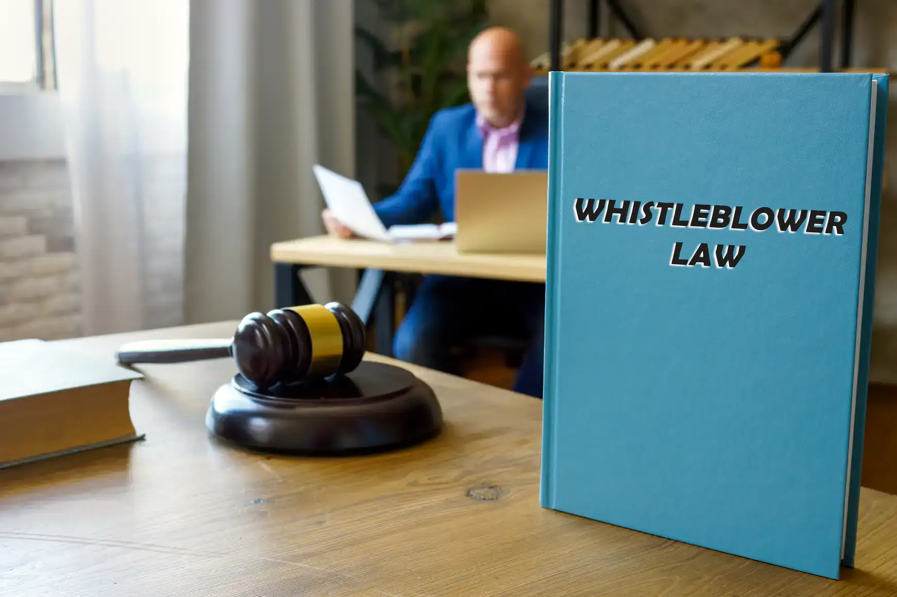 A blue book titled "Whistleblower Law" sits on a desk next to a gavel, while a sexual harassment attorney in Manhattan, NY works at a computer in the blurred background.