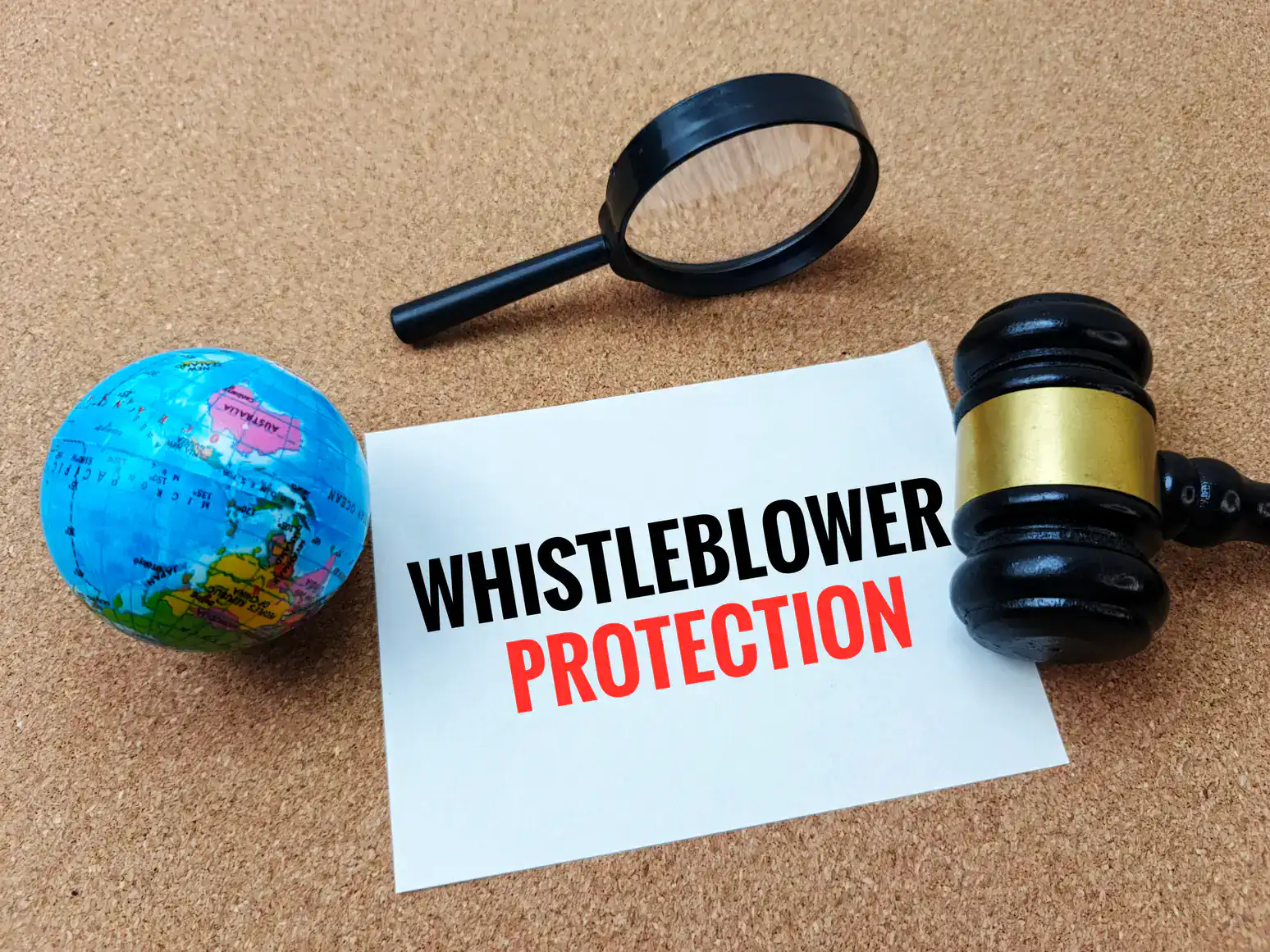 A paper with the words "WHISTLEBLOWER PROTECTION" lies on a cork surface, next to a judge's gavel, a magnifying glass, and a small globe&mdash;tools often relied upon by a sexual harassment attorney in Manhattan, NY.