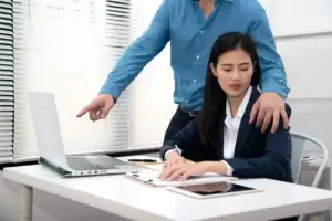 A woman sits at a desk with a laptop and tablet, looking uncomfortable as a man standing beside her places his hand on her shoulder and points at her screen—highlighting when it may be time to consult a sexual harassment attorney in Manhattan, NY.
