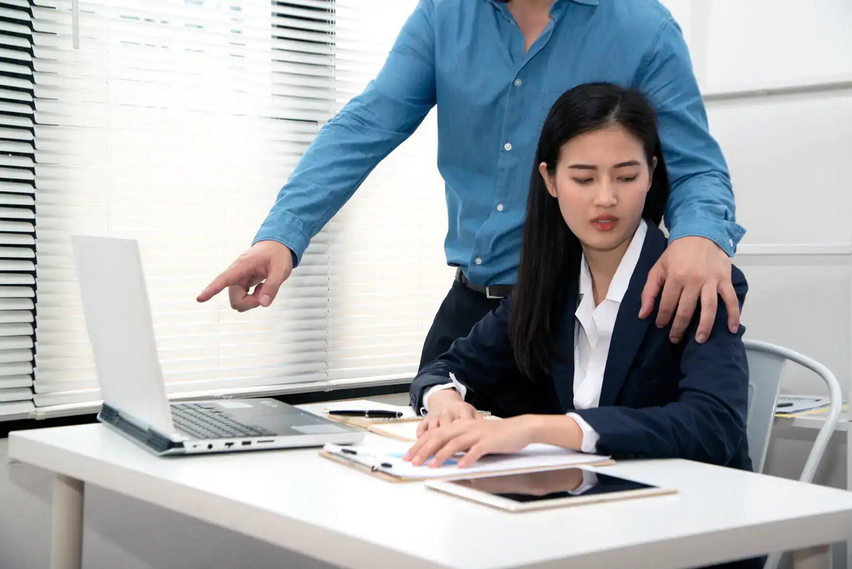 A woman sits at a desk with a laptop and tablet, looking uncomfortable as a man standing beside her places his hand on her shoulder and points at her screen—highlighting when it may be time to consult a sexual harassment attorney in Manhattan, NY.