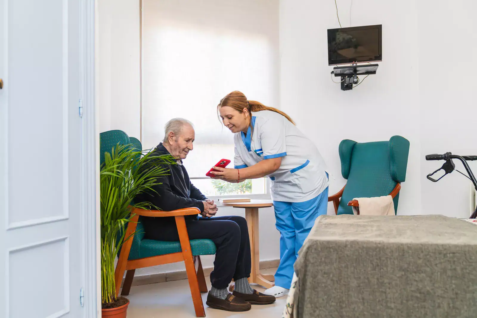 A nurse in a white and blue uniform shows a smartphone to an elderly man sitting in a cozy room with green chairs, a walker, and a wall-mounted TV—promoting awareness of nursing home fraud.