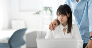 A person in a blue shirt rests a hand on the shoulder of a woman working at a laptop.