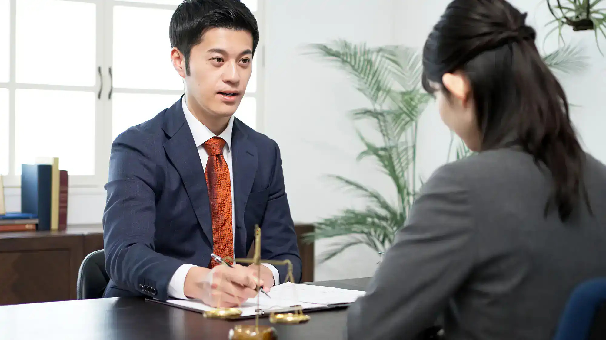 A lawyer in a suit consults with a client across a desk featuring a scale of justice.