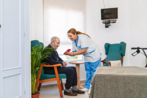 A caregiver helps an elderly man sitting in a chair by showing him a smartphone.