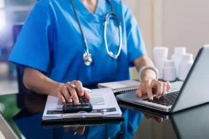 A healthcare worker in blue scrubs uses a calculator and laptop at a desk.