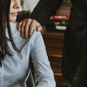 A man places his hand on a woman's shoulder as she sits at a desk, looking away. The gesture appears uninvited and the setting looks like an office.