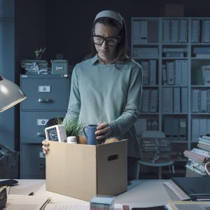 A woman in an office packs her belongings into a cardboard box, looking sad. The dimly lit, cluttered room hints she may be leaving due to wrongful termination NY, as paperwork and files are scattered around her desk.